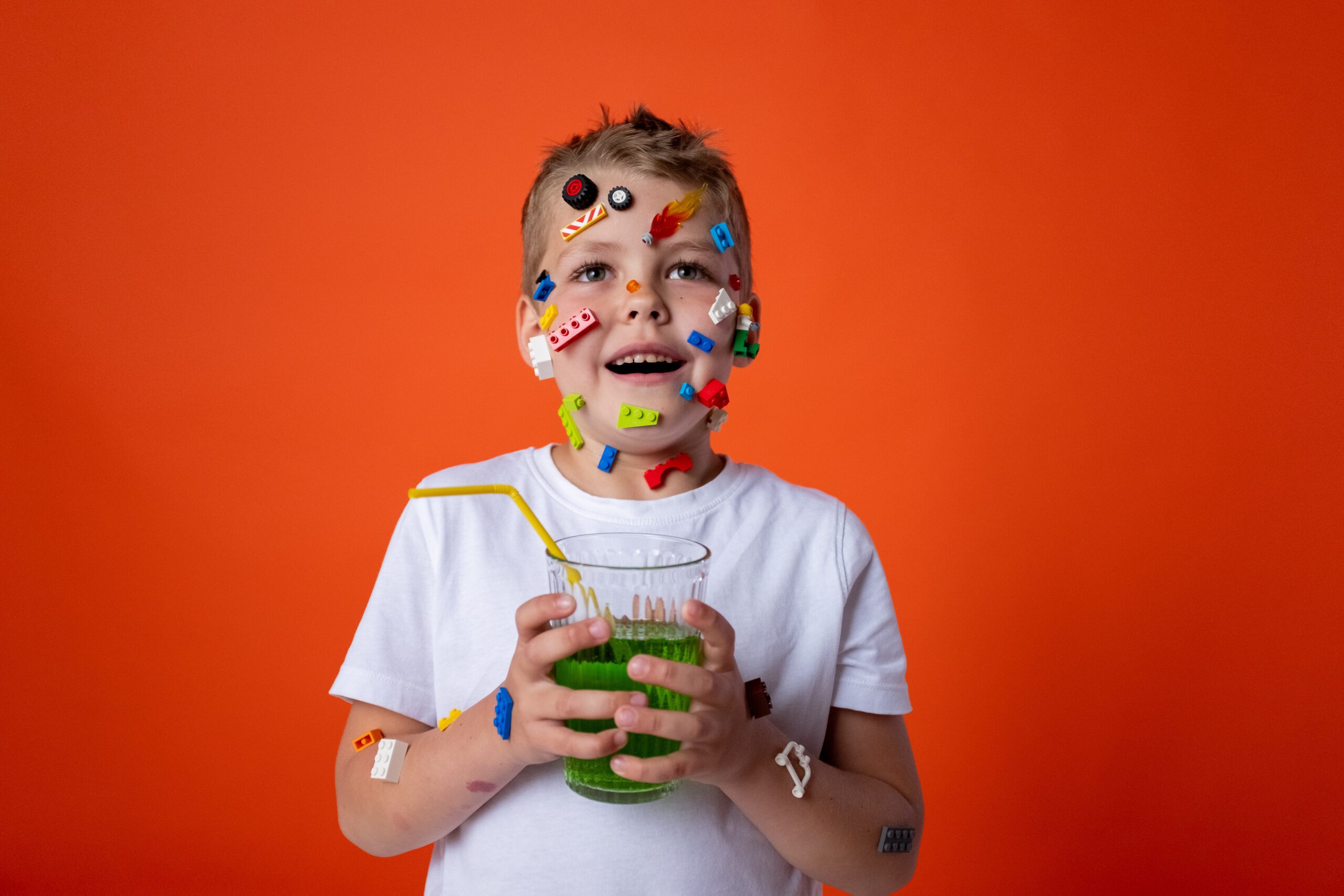 Chłopczyk trzymający szklankę z zielonym koktajlem i z przyklejonymi do twarzy elementami klocków LEGO
